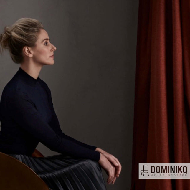 Vrouw bij een stijlvol rood gordijn van Noon, ideale dim-out gordijnstof van Kendix, De Ploeg, beschikbaar vanaf 5 meter met korting.