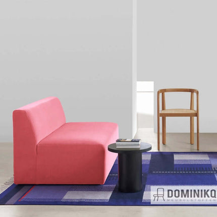 Roze bank met zachte wol van Byram-collectie, ontworpen door Raf Simons, naast een zwarte bijzettafel en houten stoel, op blauw kleed.