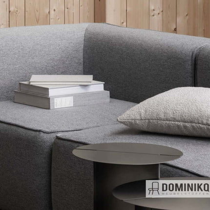 Grijze bank bekleed met Ecriture meubelstof en stapel boeken naast een kussen in een moderne woonkamer.