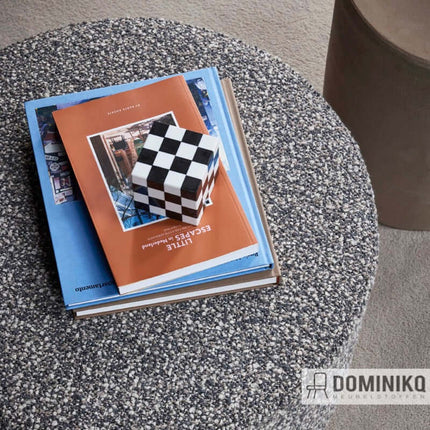 Grijze bouclé-stof van De Ploeg op een ronde tafel, met gestapelde boeken en een zwart-wit geblokt object, "Nuvola - 55".